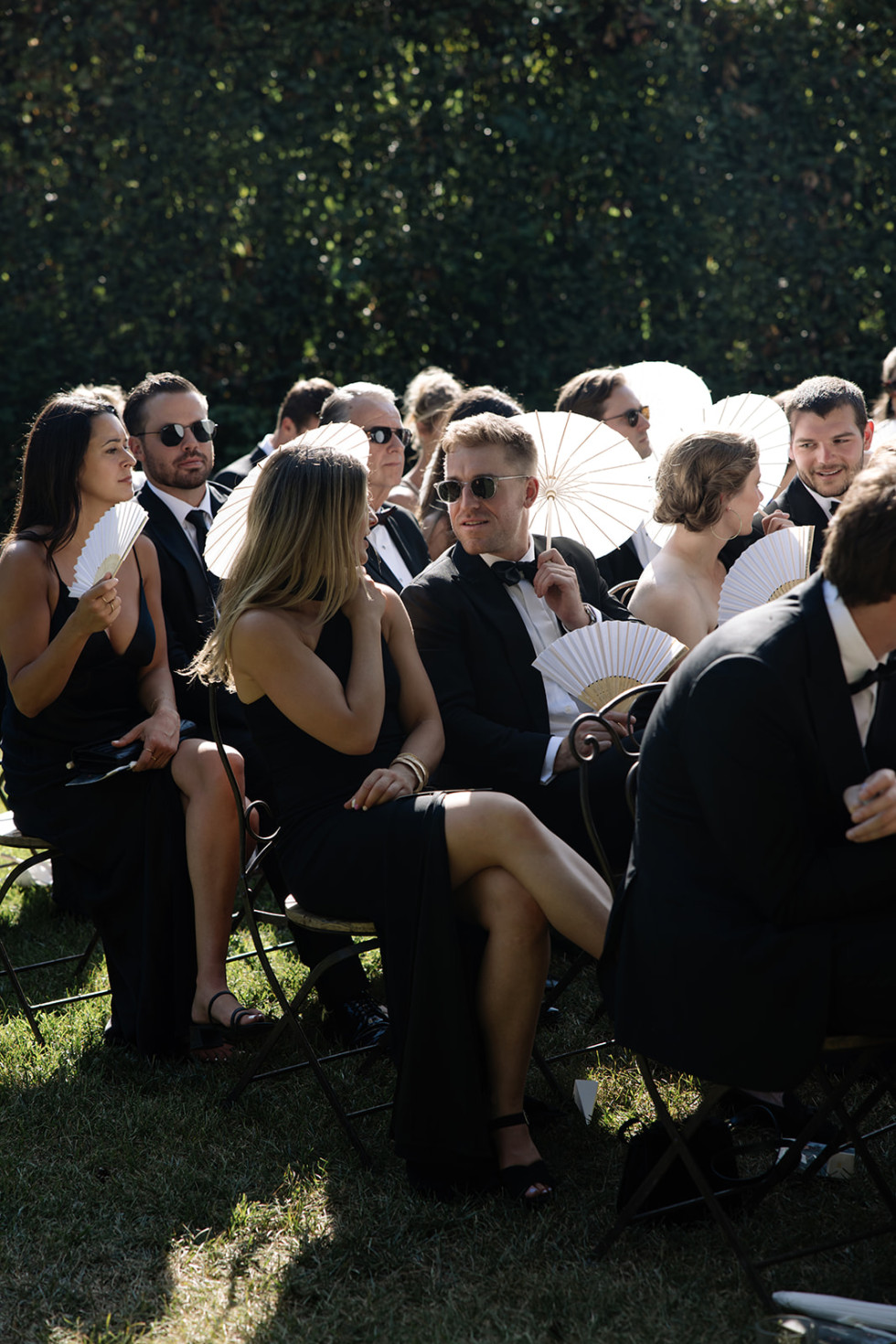 Bröllopsgäster sitter på stolar i rader och väntar på att vigseln ska börja i en solig trädgård.